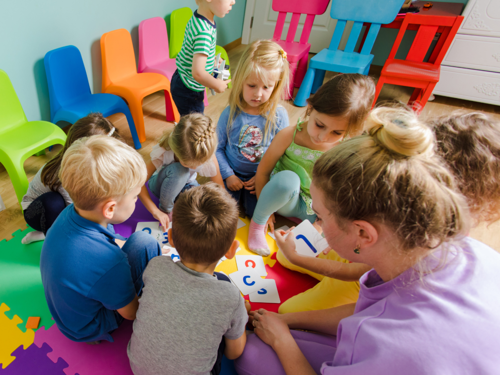 A group of approximately seven young children and a female teacher are seated in a circle on the floor, focused on an educational activity involving colorful letters or shapes. The teacher, wearing a purple top, is on the right. Brightly colored chairs (orange, pink, blue, and green) are visible in the background of the nursery or classroom.