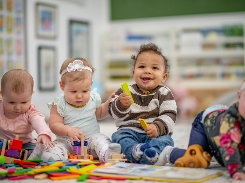 Two happy babies/toddlers, one wearing a tiara and the other wearing a striped sweater, are sitting on the floor in a bright play area, playing with blocks and colorful toys.