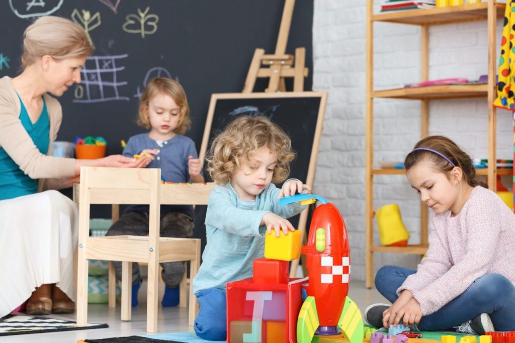 Three young children are playing in a brightly decorated classroom or playroom. A curly-haired child in a light blue shirt is sitting on the floor, stacking colorful blocks next to a large red toy rocket. Another child is sitting at a small wooden table in the background, and a third child is partially visible on the right. A blackboard with chalk drawings and an easel is behind them.