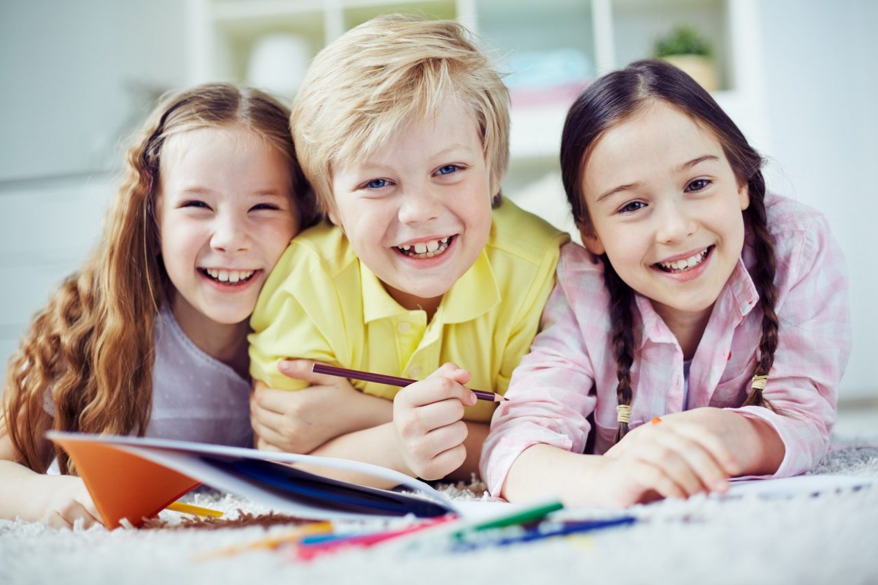 Three smiling children lying on the floor with colored pencils and paper, enjoying an art activity together.