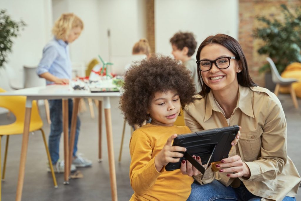 A smiling adult and a child using a tablet together in a classroom, with other children engaged in activities in the background.